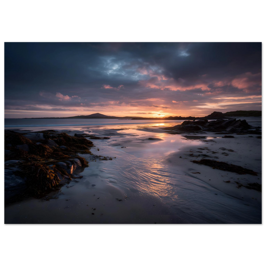 Moody violet and gold sunset over an empty beach at low tide, seaweed-covered rocks and a tidal channel across wet sand, in a slim dark frame