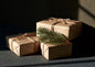 Three gifts wrapped in brown kraft paper and tied with jute twine, a sprig of spruce under one knot, lit from one side against a dark ground
