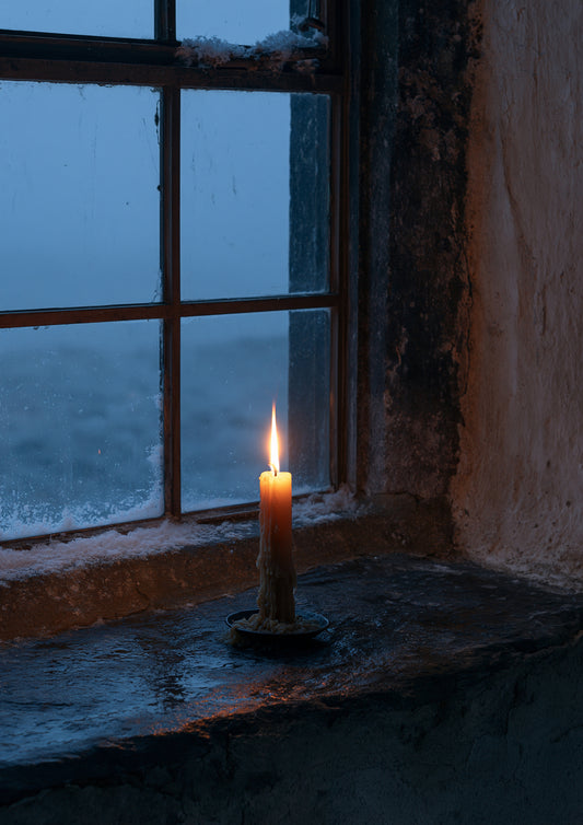 A single lit candle on a deep stone windowsill seen from inside a dark room, snow falling in the blue dusk beyond the glass