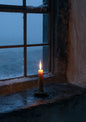 A single lit candle on a deep stone windowsill seen from inside a dark room, snow falling in the blue dusk beyond the glass