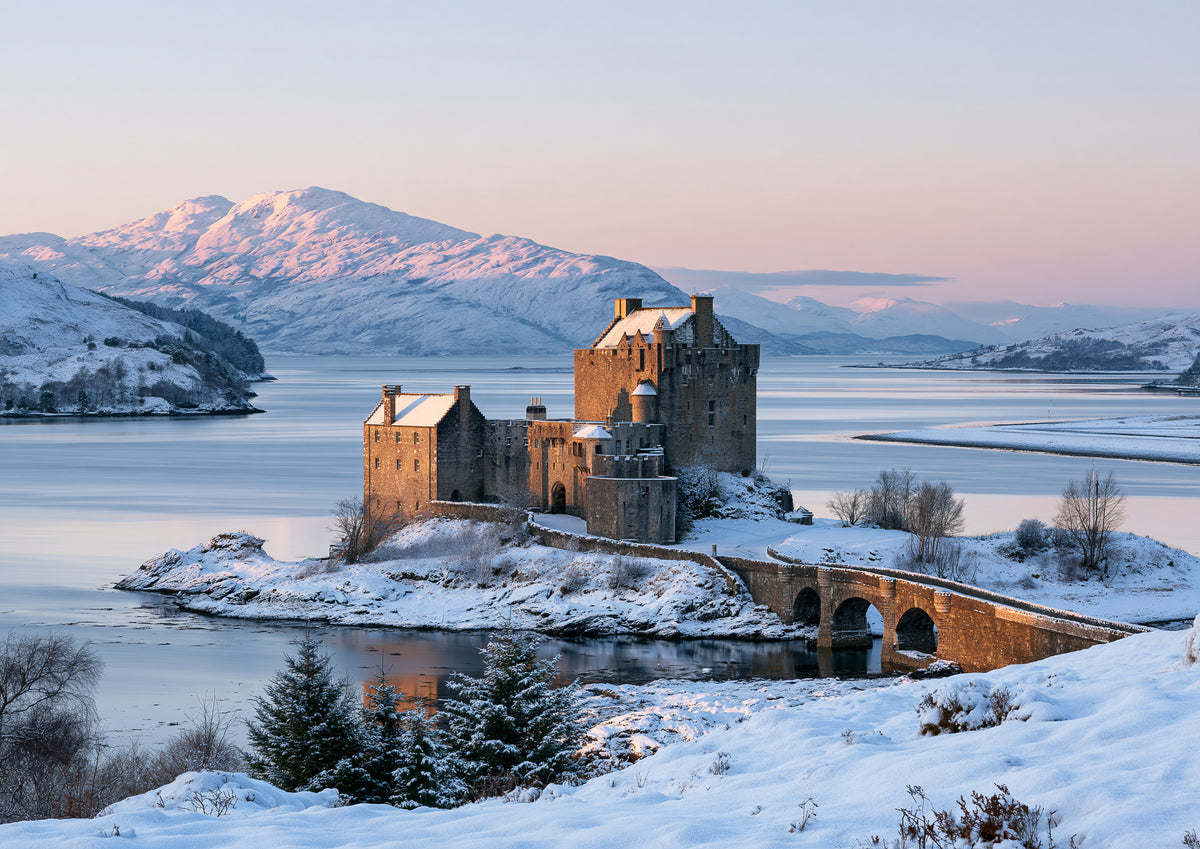 Eilean Donan Castle on its snowy tidal island at winter dawn, the three-arched stone bridge leading out to it and snow-capped mountains glowing pink beyond the loch