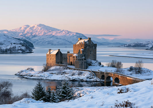 Eilean Donan Castle on its snowy tidal island at winter dawn, the three-arched stone bridge leading out to it and snow-capped mountains glowing pink beyond the loch