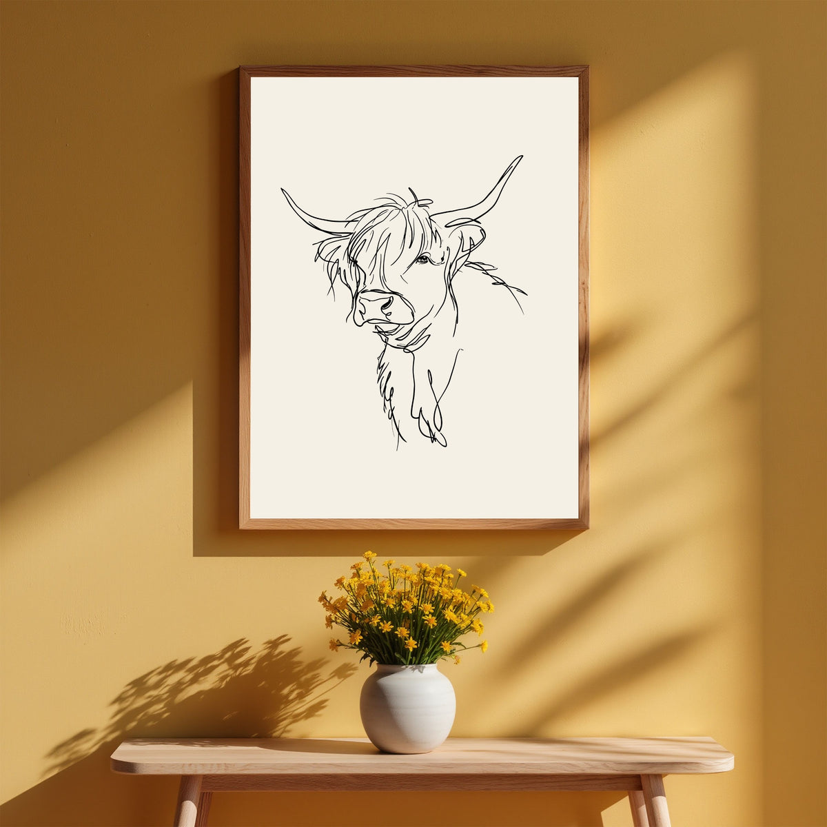 Loose line sketch of a Highland cow, in a light wood frame on a warm ochre wall above a bench with a vase of yellow flowers