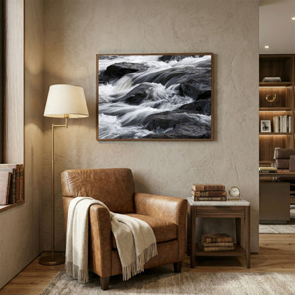Silk water over dark rock, in a pale wood frame above a tan leather armchair in a book-lined reading corner