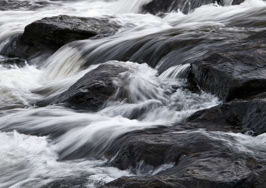 Pale water pouring over near-black wet rock, the flow smoothed into long soft streaks like silk laid over stone