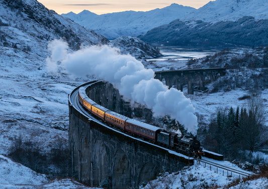 A steam locomotive crossing a long curved stone viaduct over a snowbound Highland glen at winter dusk, a white plume of steam trailing behind