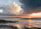 A towering storm cloud lit cream and apricot by a setting sun above a quiet bay, a slate-dark front sweeping in overhead and wet sand at low tide reflecting the sunset