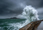 Storm wave bursting white over an old stone sea wall, dark green sea and an island on a grey horizon