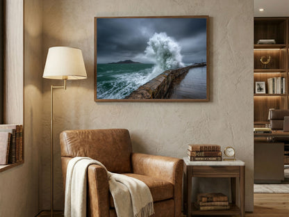 Storm wave bursting over a stone sea wall, framed above a leather armchair in a book-lined study