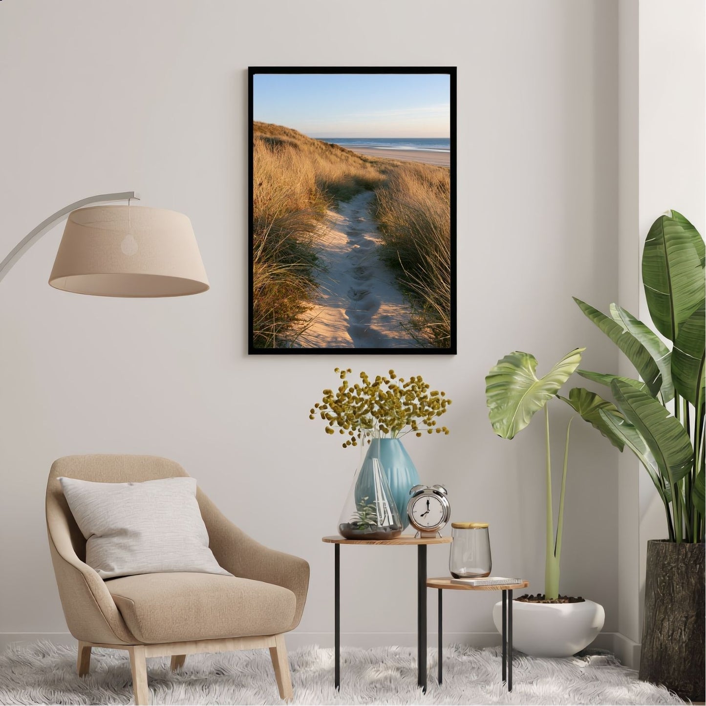 Sand track through marram grass to a beach, in a black frame above an armchair in a bright reading corner
