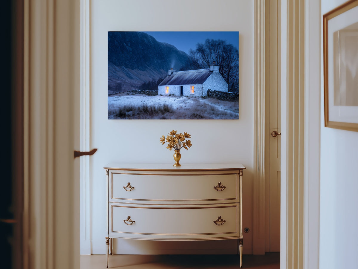 Lit croft house in snow at blue hour, hanging above a painted chest of drawers in a bright hallway