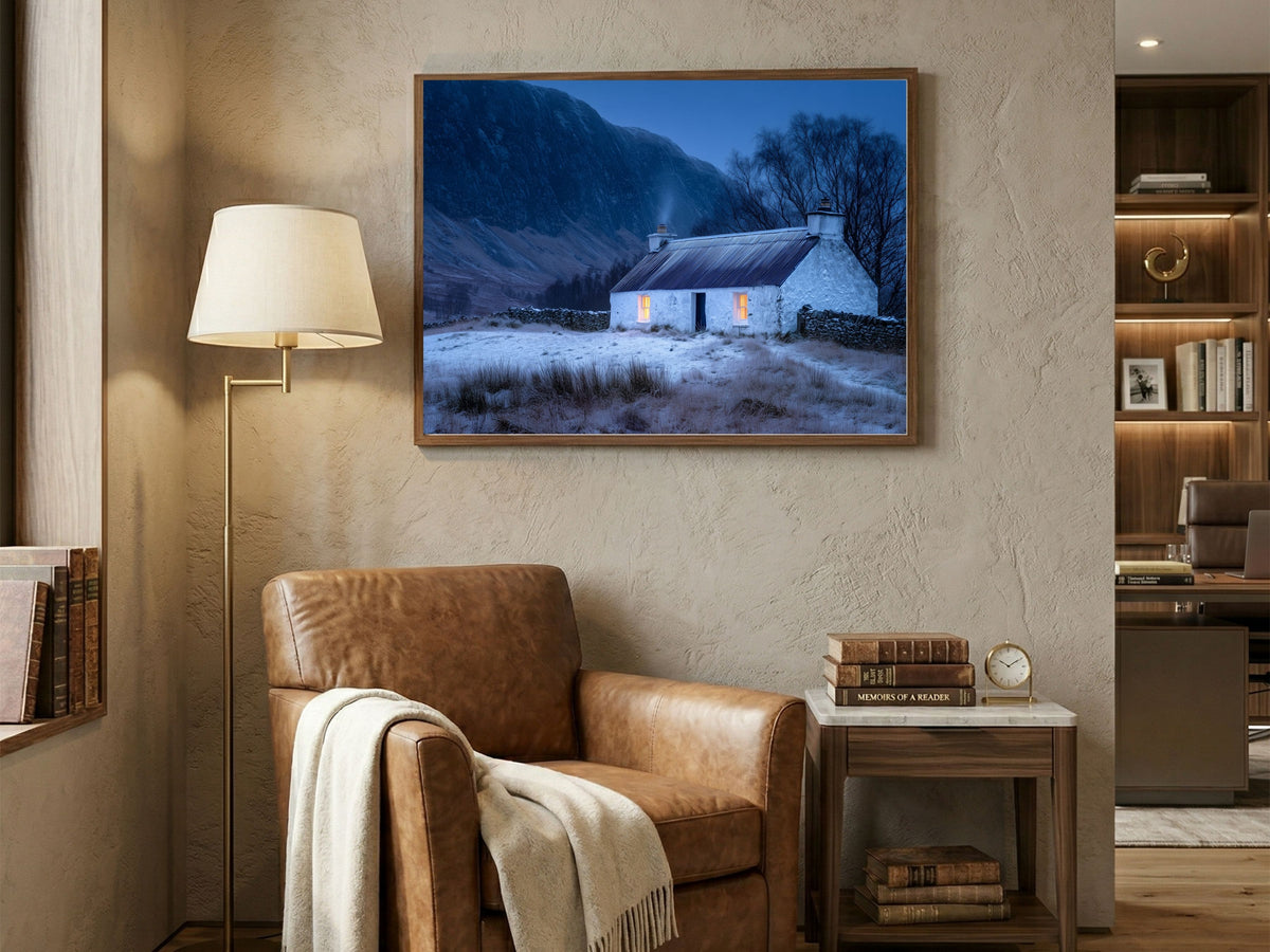 Lit croft house in snow at blue hour, in a wooden frame above a leather armchair in a warm living room